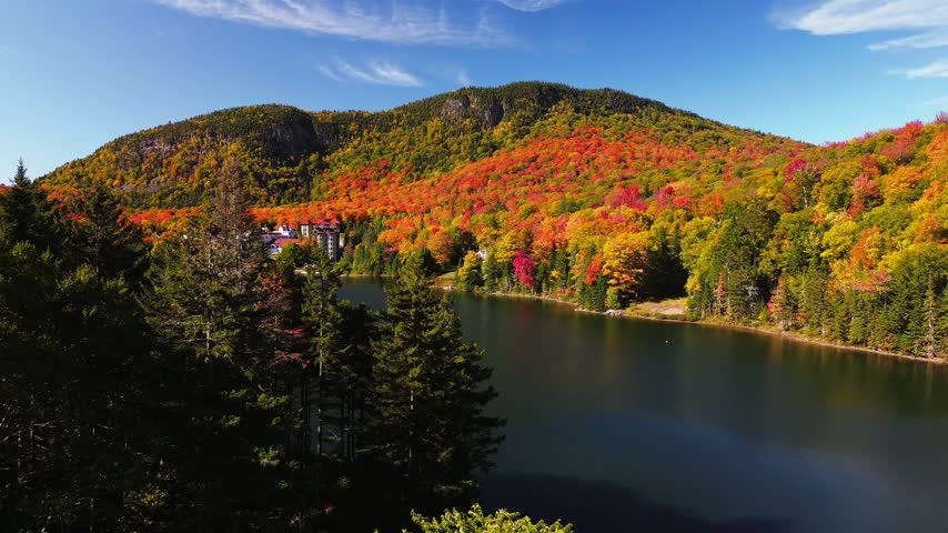 Sweeping aerial footage of a picturesque Vermont lake nestled among forested mountains ablaze with autumn color. Brilliant fall foliage blankets the surrounding hillsides, while deep blue water winds past a lakeside resort beneath a clear autumn sky. This scenic overhead view captures the beauty of New England during peak fall season, ideal for travel promotions, destination marketing, seasonal nature films, and scenic landscape sequences.
