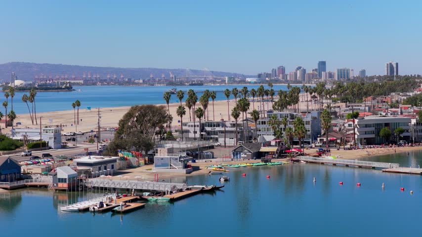 Colorful kayaks line a sheltered sandy beach on Alamitos Bay in Long Beach, California. Floating docks, red buoys, palm trees and beachgoers. Downtown skyline, port cranes and bridge on a sunny day.