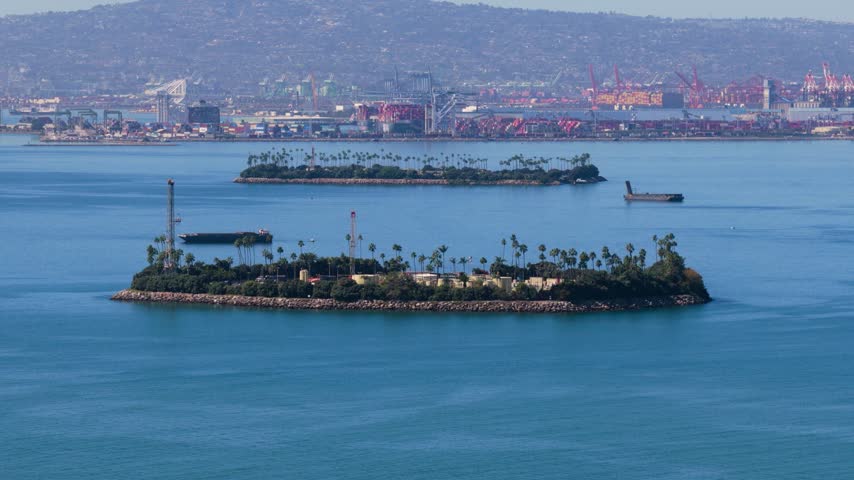 Artificial oil drilling islands with derricks, palm trees and rock shores in Long Beach harbor, California. Barges float on blue water before red port cranes, container stacks and hazy hills.