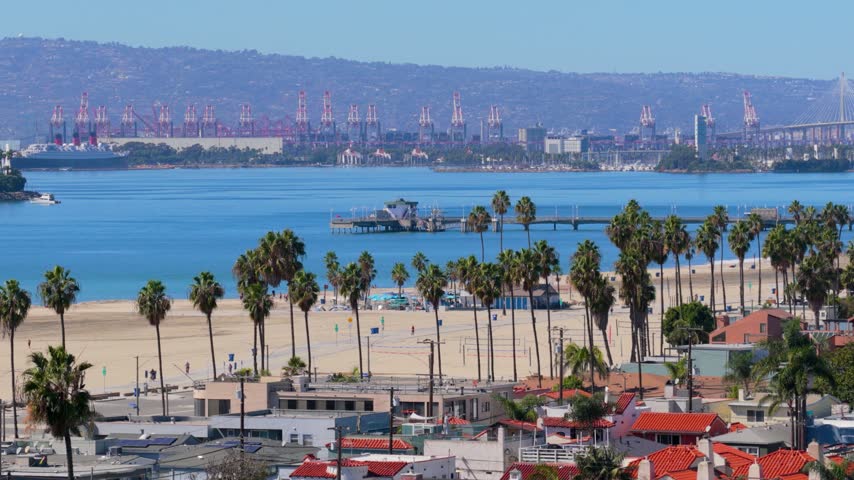 Belmont Pier stretches into the bay at Long Beach, California, with the Queen Mary, red port cranes and a cable bridge beyond. Palm trees and volleyball nets line a sunny beach above red tile roofs.
