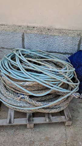 Coiled blue thick nautical ropes and blue commercial fishing nets stacked on a wooden pallet and quay at a maritime port in Spain.