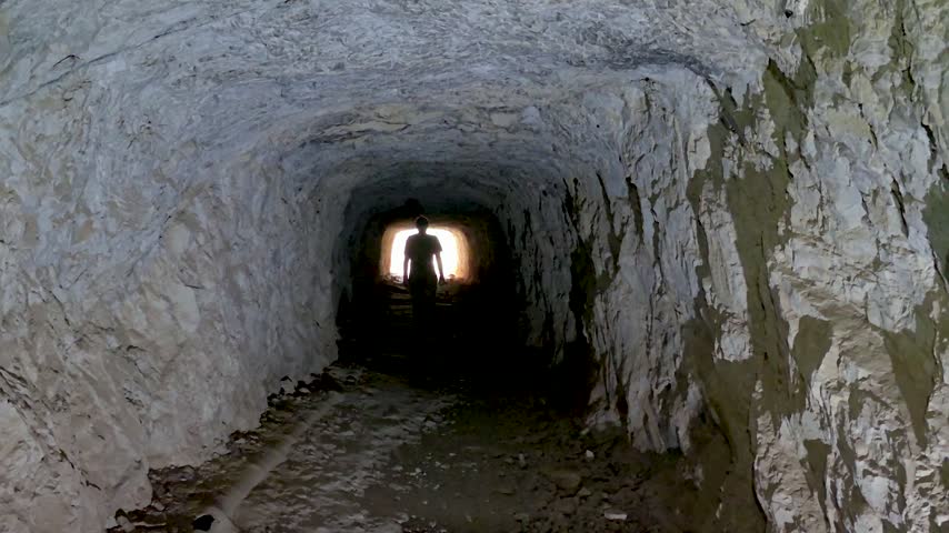 Silhouette of a person walking inside a dark rock tunnel towards a bright glowing exit light, conceptual footage of hope, way out, future and achievement.