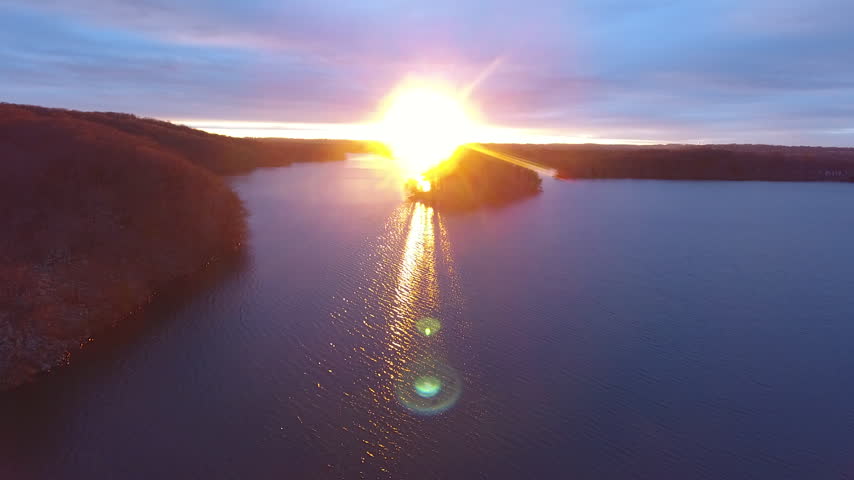 Aerial drone shot of a bright setting sun with lens flare over a wooded island and calm water at Loch Raven Reservoir under a purple sky. Baltimore County, Maryland, USA.