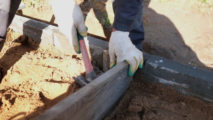 Gloved construction workers align wooden formwork and compact soil beside concrete edging on sunny outdoor site, showing hands-on groundwork, careful leveling, and building progress.