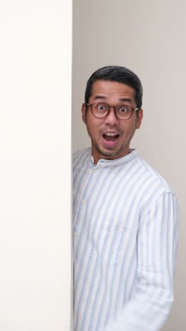 Adult Asian man peeking from behind a wall cheering happy to celebrate something