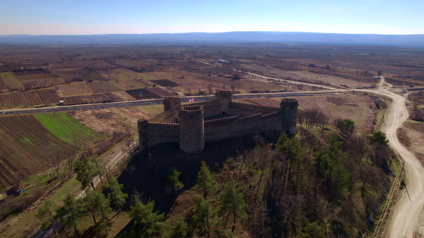 Rectangular stone fortress fills more of the frame as the drone approaches from above fields revealing crenellated walls four round corner towers a courtyard and the paved road behind the site