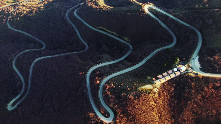 Hairpin road bends cross a wooded hillside as the aerial view widens and shifts right bringing a lower loop and small roadside buildings toward the edge of the frame under slanting sunlight