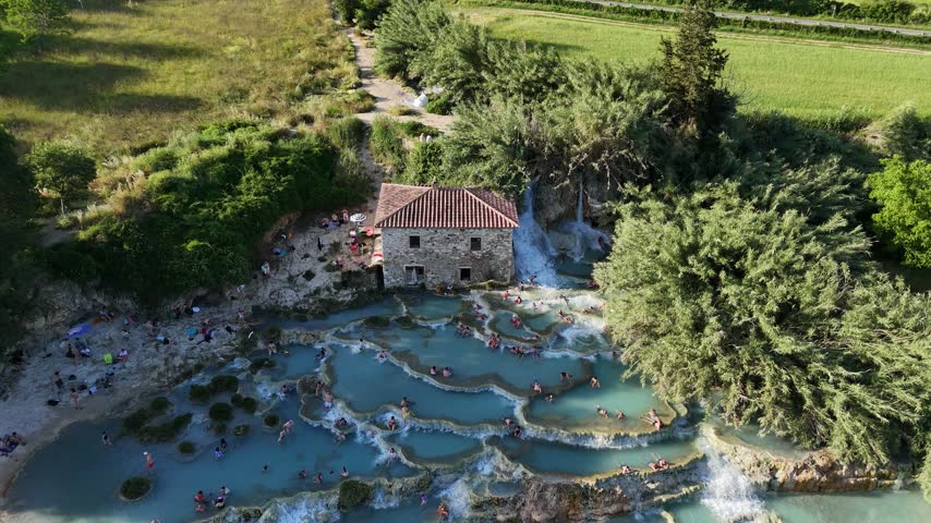 Cinematic aerial orbit around the historic stone mill house standing beside the natural thermal waterfalls of Saturnia.
