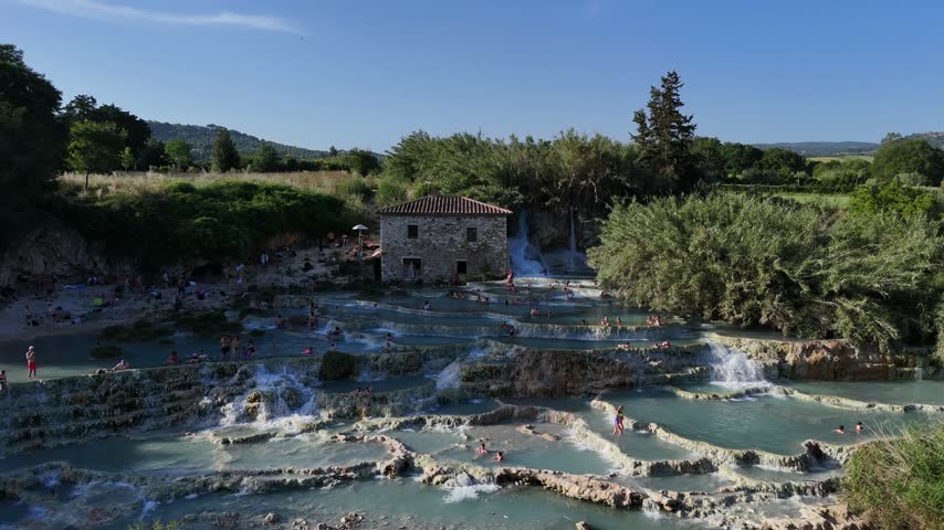 Eye-level aerial shot facing the iconic rustic mill house behind the steaming turquoise travertine pools of Saturnia.
