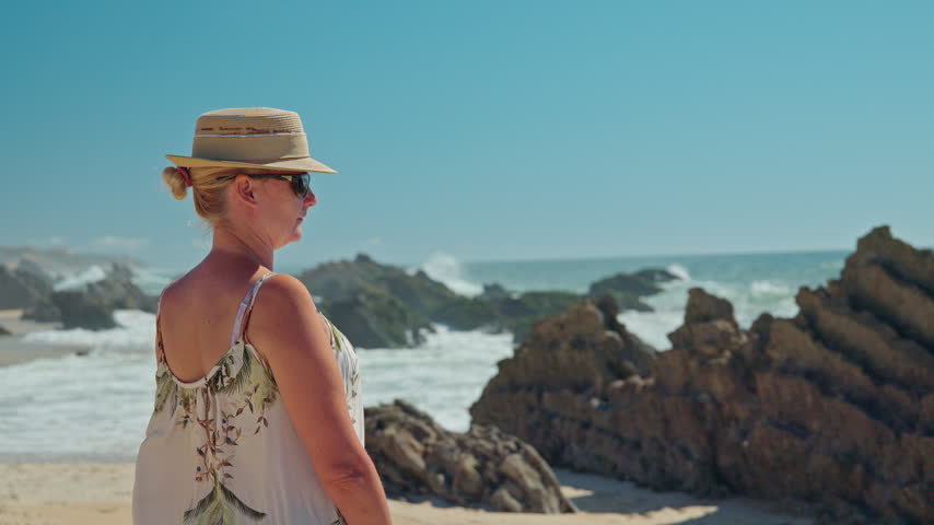 Woman in a straw hat watches waves breaking against rocky shoreline on a sunny Portuguese beach.