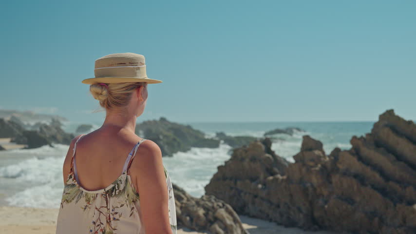 Woman in straw hat gazes at ocean waves breaking around rocky formations on a sunny Portuguese beach.