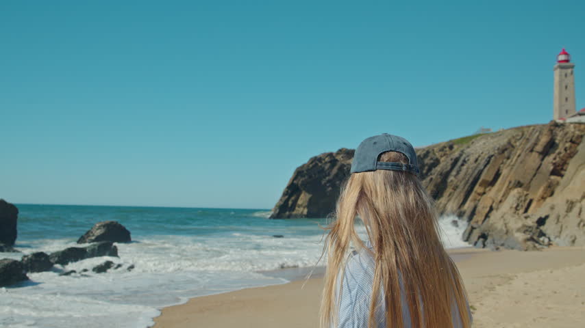 A young woman in a cap watches waves breaking on a sandy Portuguese beach below a cliffside lighthouse.