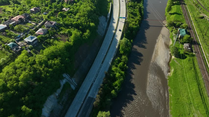 Drone view of a modern highway bridge running alongside a river in the Georgian countryside. The scene features green vegetation, riverside homes, agricultural land, and a winding waterway