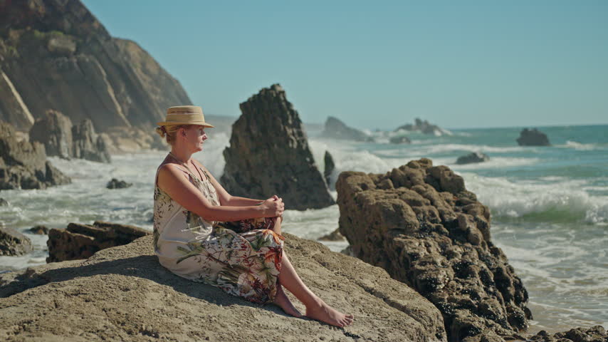 A woman in a straw hat sits on coastal rocks beside ocean waves in Figueira da Foz, Portugal.