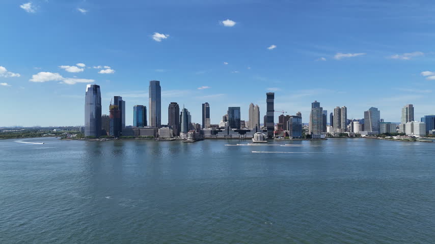 Aerial pan across the Hudson River from Lower Manhattan toward the Jersey City skyline and waterfront under a blue sky with clouds, New York, USA.