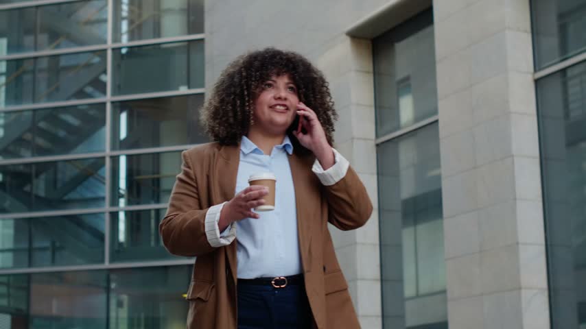 Confident businesswoman with curly hair making a phone call and holding coffee outdoors