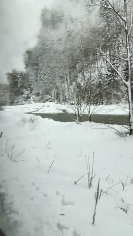 A moving train window view follows a river through a forest covered in winter snow.