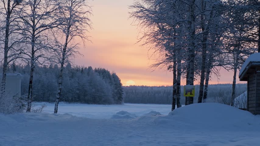 Scenic view of a glowing winter sunset casting a warm orange light over a frozen lake, snow covered ground, and a dense pine forest in Lapland Finland