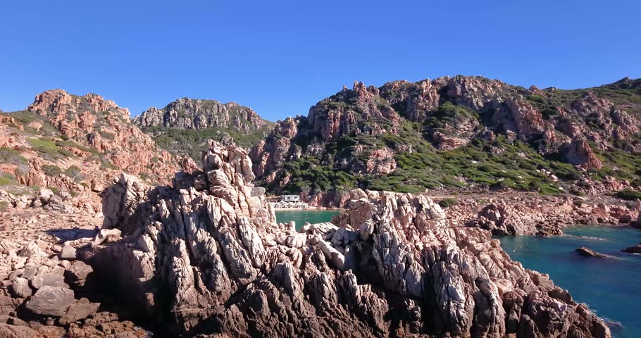 Aerial approach to a sheltered sandy beach framed by rugged granite cliffs and emerald water at Cala Li Cossi, Sardinia.