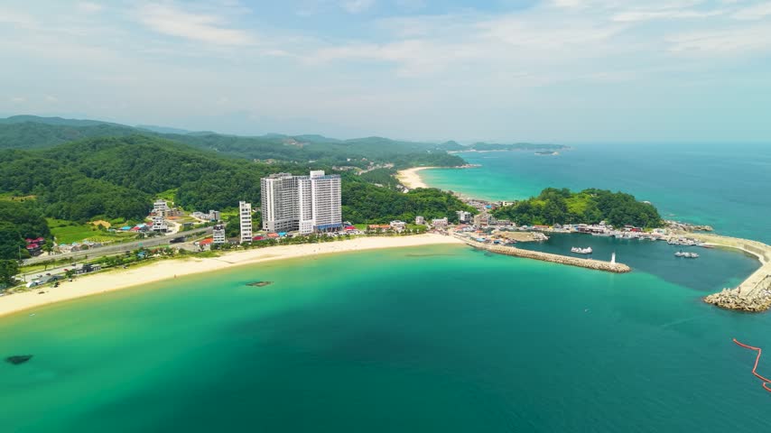 Distant aerial view of Ingu Beach and Jukdo Beach with turquoise bay, high rise resort buildings, fishing harbor and green forested hills along coast in Yangyang, Gangwon Province, South Korea
