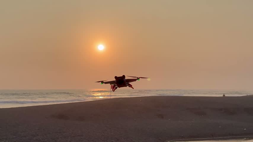 Drone flying low over a sandy beach at sunset, silhouetted against a warm orange sky with the sun reflecting on the ocean waves