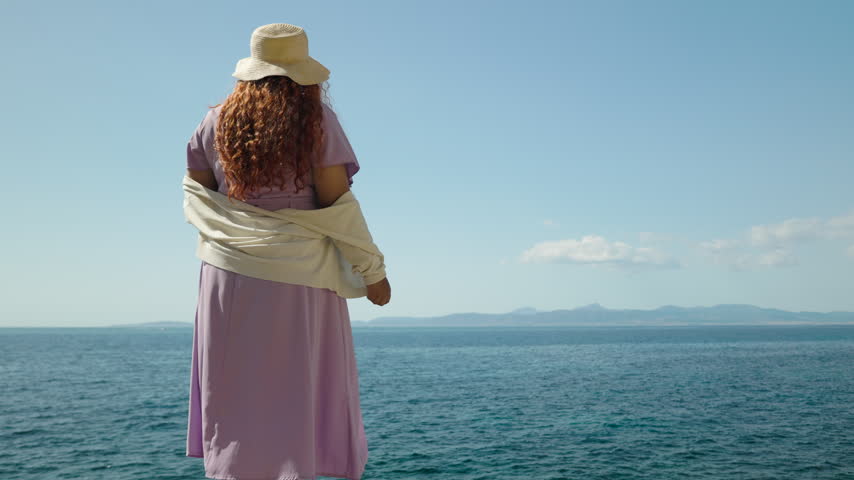 Red haired woman seen from behind looking at the Mediterranean Sea horizon on a sunny summer day. Coastal landscape with ample space for text, vacation and travel concept