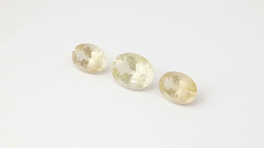 Brilliant light reflects across multiple polished facets of transparent oval rutilated quartz gemstones rotating smoothly on a neutral background revealing intricate internal rutile needle inclusions