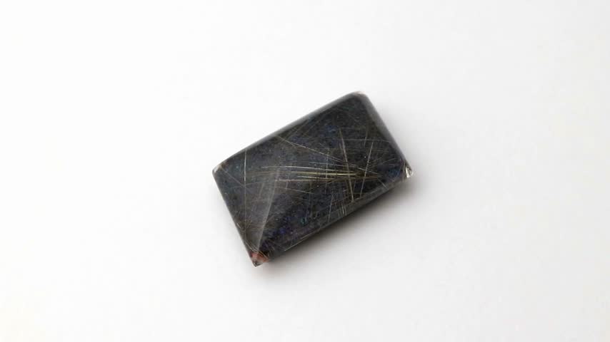 Polished rectangular triplet gemstone displays intricate golden needle inclusions and subtle iridescent color flashes across a dark opaque surface rotating smoothly on a clean background.
