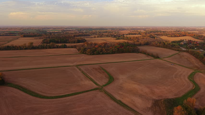 Aerial establishing shot of broad agricultural fields divided by green strips and wooded patches, with a winding road and scattered houses visible beyond the fields in northern Wisconsin