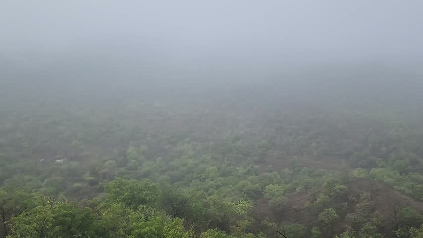 Aerial view of a misty mountain valley covered in a dense forest during the monsoon season with heavy fog rolling over the hills.