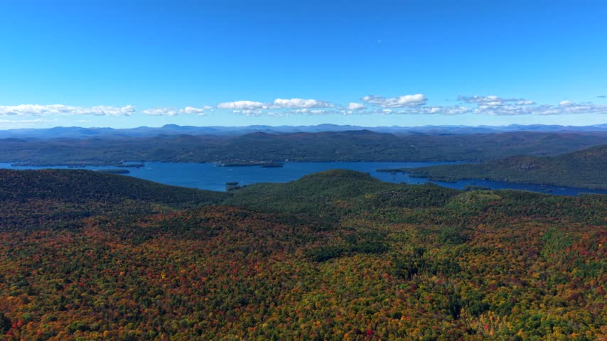 Sweeping panorama from Sleeping Beauty Mountain overlooks Lake George, fiery autumn canopy spreading toward Adirondack ridgelines beneath blue sky with scattered clouds. Aerial drone wide shot