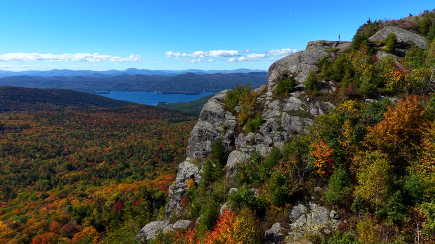 Hiker stands on cliff edge above Sleeping Beauty Mountain, Lake George shimmering below through vivid autumn canopy, hazy Adirondack ridgelines fading beneath blue sky. Aerial drone forward shot