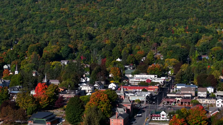 Telephoto descent over Lake George, Adirondacks, New York, compressing a waterfront village of brick buildings and marinas against a hillside of autumn color
