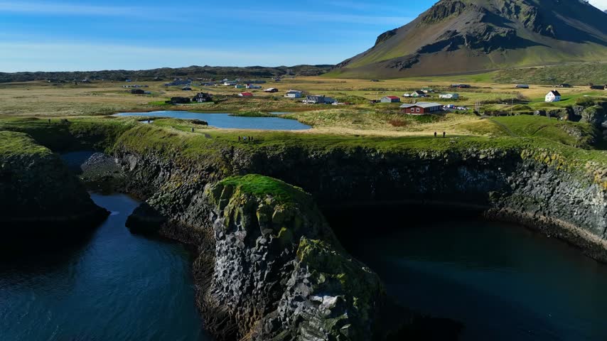 Aerial view over Arnarstapi, Iceland, with rugged basalt cliffs, a small pond, scattered homes and a dramatic volcanic mountain rising beyond the grassy coast.
