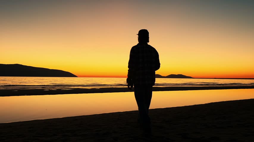 A dark profile of a man wearing a baseball cap stands along the shoreline, raising a bottle to his mouth. Vibrant amber skies, distant islands and reflections on wet sand frame this quiet summer evening moment.