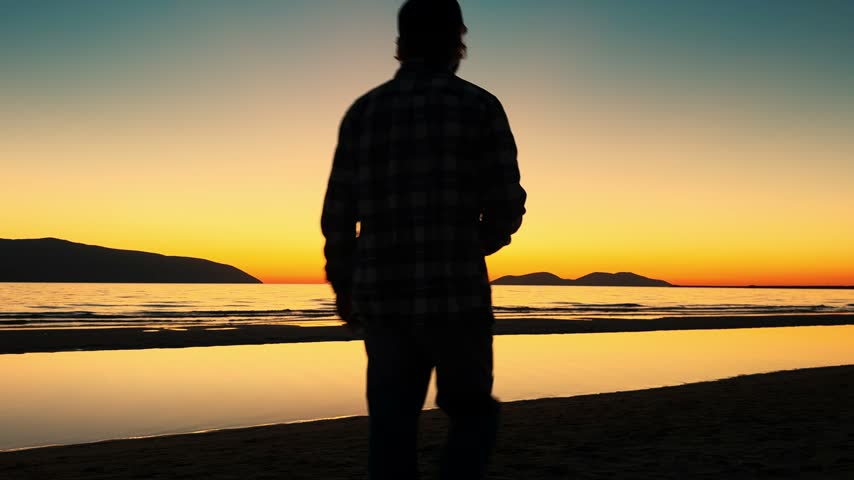 A dark profile of a man wearing a baseball cap along the shoreline, raising a bottle to his mouth. Vibrant amber skies, distant islands and reflections on wet sand frame this quiet summer evening moment.