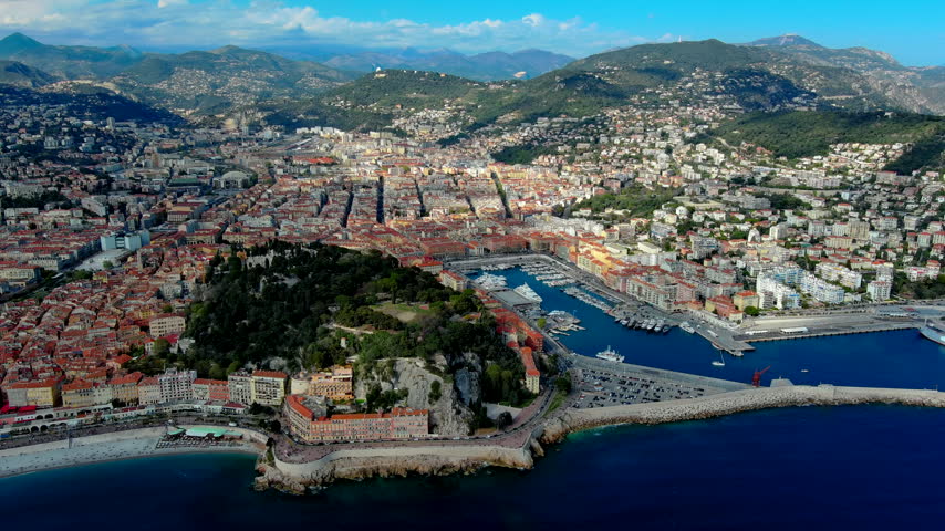 High-angle aerial panorama of Nice, France, with Port Lympia, Castle Hill, yachts and sailboats, colorful historic buildings and mountain foothills beside the Mediterranean.