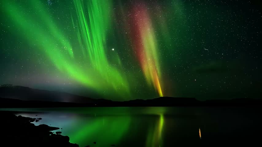 Vivid green and red northern lights shining over a still lake that mirrors the glow, with dark hills and a sky full of stars above the shoreline