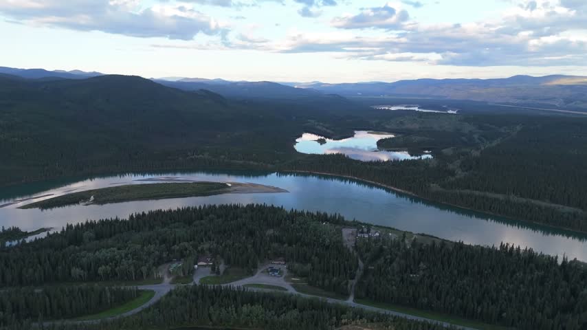 Drone flyover of a slow river and small shining lakes in a dark spruce valley, with a few scattered homes and driveways below layered hills at dusk