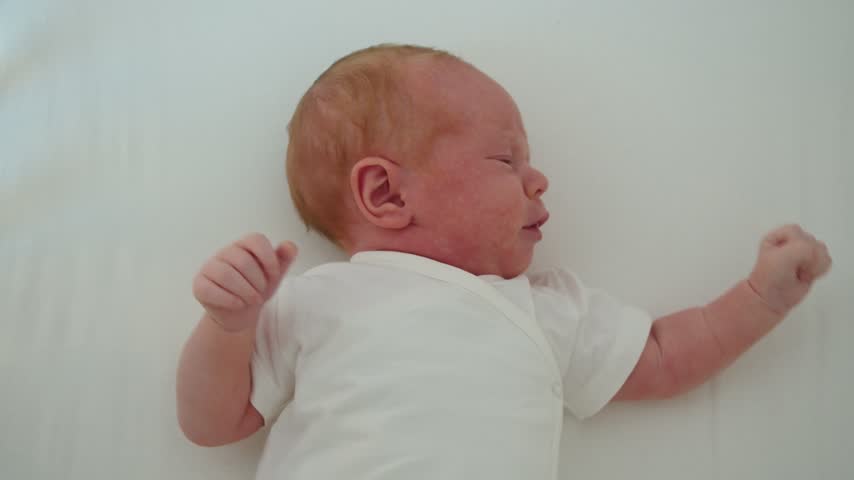Newborn lies in crib with noticeably red skin tone, moving gently and showing delicate appearance, early adaptation, and natural newborn features during quiet home care in nursery indoors.