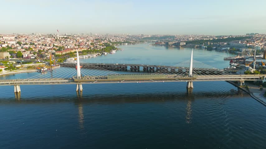 Istanbul, Turkey. The Golden Horn Metro Bridge spans the Halic estuary in warm morning sunlight, its twin white pylons and harp-arranged cables forming a striking cable-stayed structure. The bridge carries the M2 metro line and features the unusual Halic Metro Station integrated directly into its central span, a design that places platforms above the water between the Beyoglu and Fatih districts. An aerial drone perspective reveals the bridge