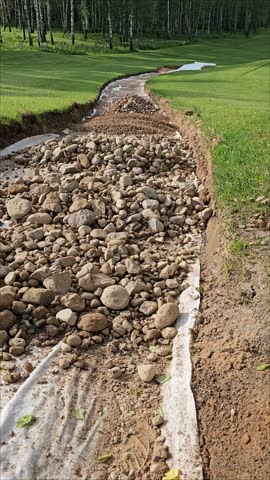 A rock-lined drainage channel curves across grass in a park bordered by trees, with pale banks and stones visible in daylight.