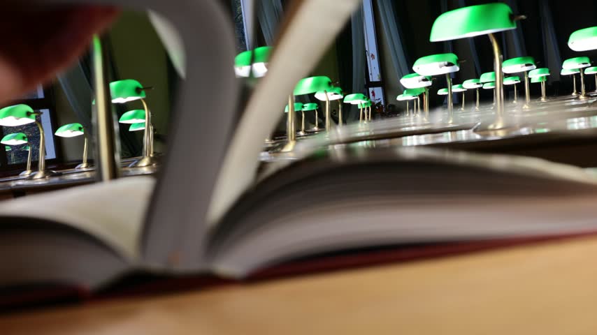 Hand turning pages of open book in library with rows of green banker lamps behind
