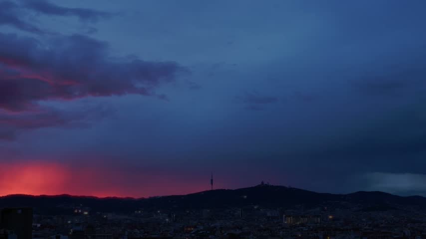Dark clouds shifting over barcelona skyline at dusk, revealing colorful twilight glow behind mountains