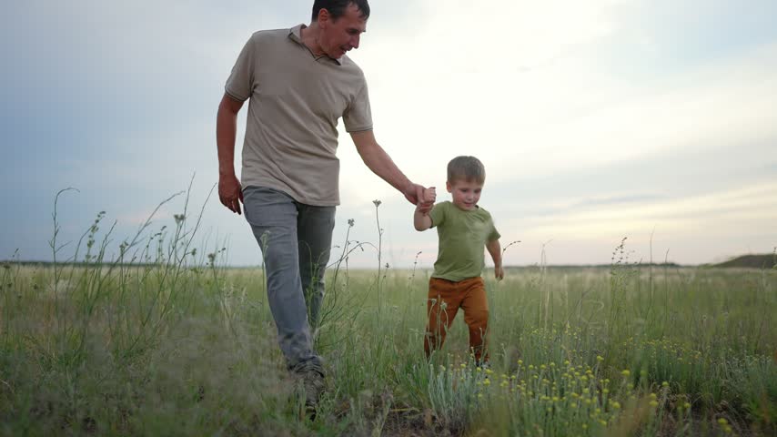 Father and son walk together holding hands in grassy field at sunset. Happy child smiles while walking beside dad outdoors. Father holds child hand in nature. Family bonding in summer field.