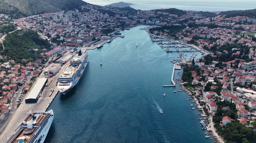 Aerial view of Dubrovnik Bay, Croatia, with tourist and cruise ships sailing through blue Adriatic waters, surrounded by a dramatic coastline, green hills and scenic landscapes.