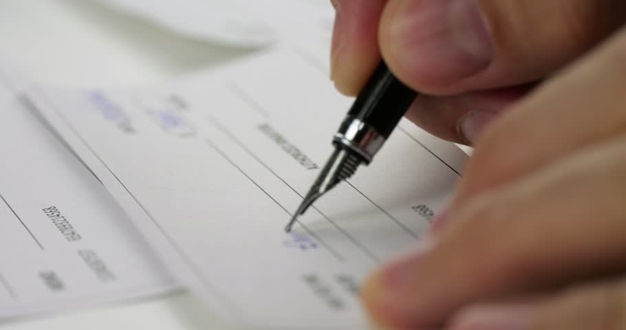 Close-up of hand signing financial document with black pen. Person completes paperwork, check, or payment form on desk. Slow writing shows business, banking, finance, and office administration.