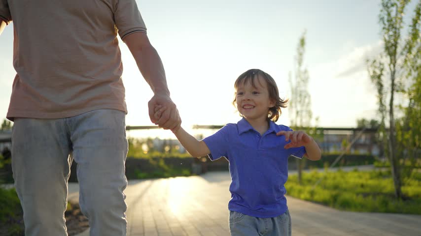 Father and son walk together holding hands in park at sunset. Happy boy looks up smiling at dad during evening stroll. Child holds hand of father on pathway. Family enjoys walk in warm sunlight.