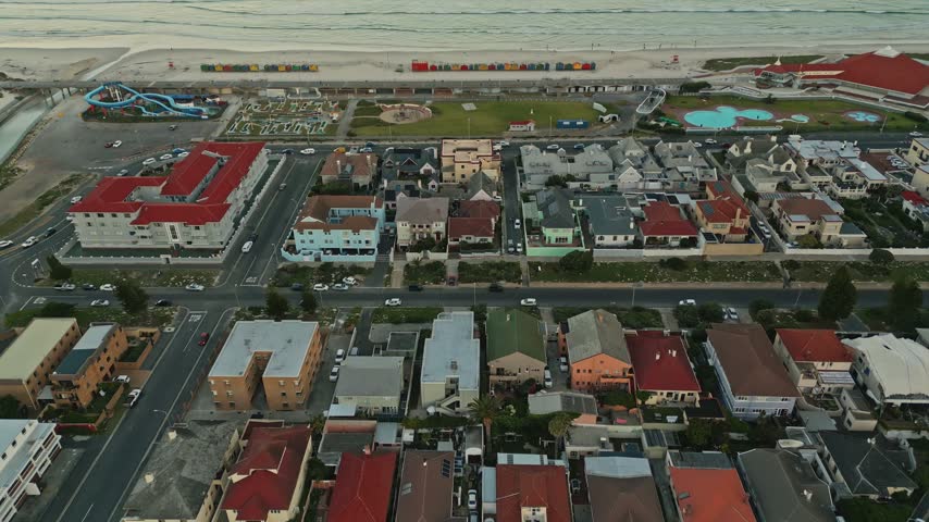 Colorful Muizenberg beach huts line sandy coast beside ocean waves, while aerial golden hour view highlights bright facades and warm sunset atmosphere in Cape Town, South Africa, during sunset.