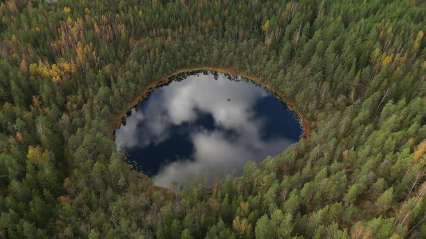 Panoramic drone footage of a circular lake in autumn woodland with glassy water reflecting the open sky and surrounding forest